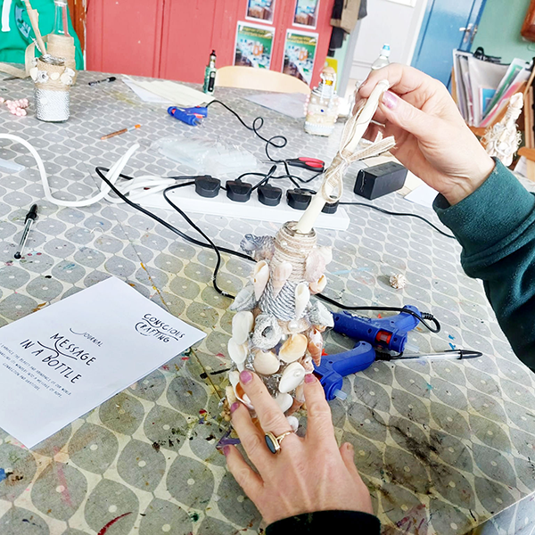 Person decorating bottle with seashells, rope, and ribbon at craft table labeled 'Coastal Curios Message in a Bottle'.
