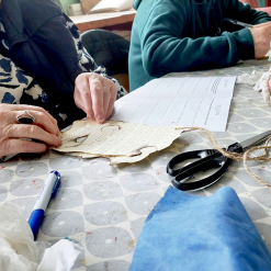 Two people working with aged paper and worksheet labeled 'CEEA' on patterned table with craft tools.
