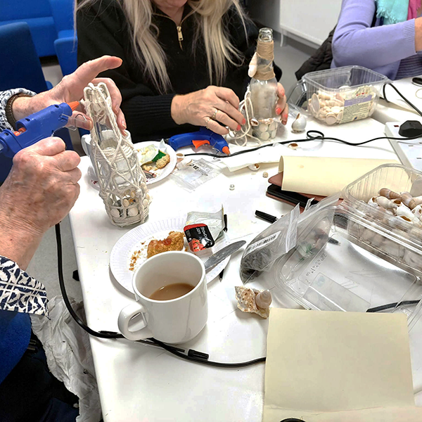 Group decorating glass bottles with seashells and string at craft table with tools and snacks.