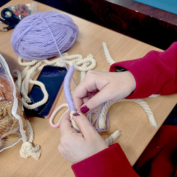 Close-up of a person wrapping light purple yarn around thick cream-colored rope at a wooden table; crafting materials include a large ball of purple yarn, pink-handled scissors, a smartphone, and a mesh bag with assorted supplies