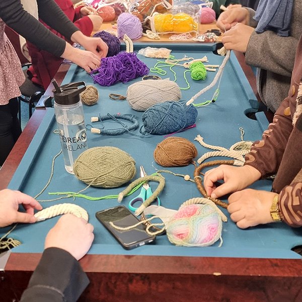 Group of people gathered around a pool table repurposed as a crafting workspace, covered with colorful yarn balls, thick ropes, scissors, and a smartphone; participants actively engaged in a yarn-based activity such as macramé, with a water bottle labeled 'ÉIREANN IRELAND' visible