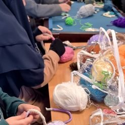 Group of individuals seated around a table engaged in a yarn craft activity, using crochet hooks and colorful yarns; a clear plastic bag filled with multicolored yarn balls is prominently placed on the table in a workshop or class setting