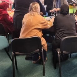 Group of people seated around a table engaged in a communal crafting activity with yarn and tools; indoor setting with chairs, a colorful mural in the background, and a collaborative, creative atmosphere