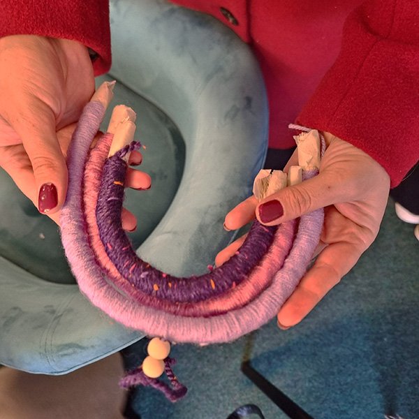 Person holding a handmade rainbow-shaped textile crafted from thick curved cords wrapped in pink and purple yarn, with frayed ends, beads, and tassels; wearing a red coat with painted fingernails, seated near a light blue cushion on a carpeted floor