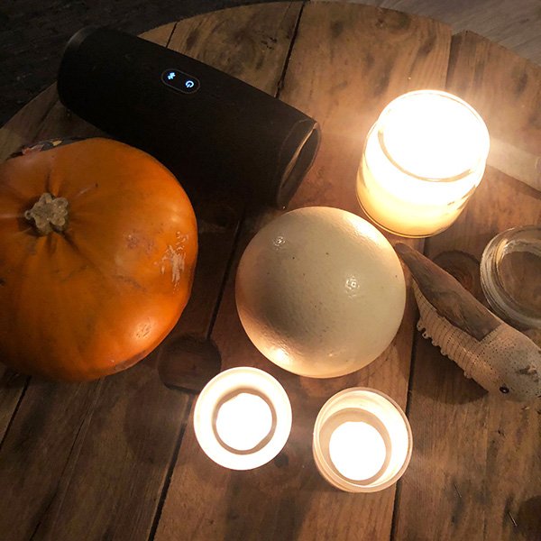 Wooden table with pumpkin, ostrich egg, lit candles, speaker, and carved fish in warm candlelight.