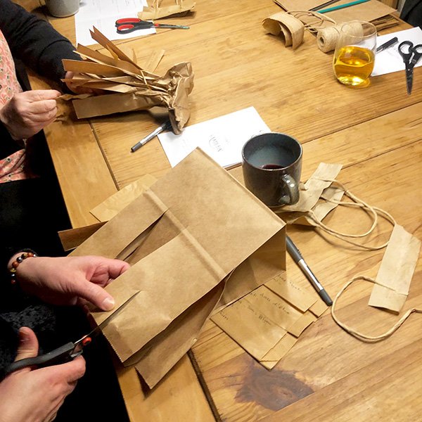 People cutting and shaping brown paper bags at table with scissors, notes, and mugs during craft session.
