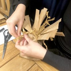 Person twisting brown paper strips into bouquet-like shape on table with scissors and white paper.