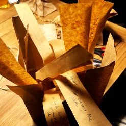 Fan-like arrangement of brown paper strips with handwritten messages on wooden table with craft tools.