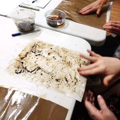 People staining paper with coffee or tea at a table with dark liquid containers and abstract sheets.