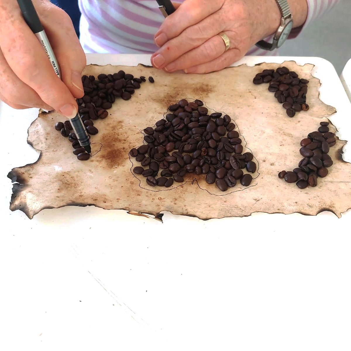 Person arranging coffee beans on aged paper, possibly forming shapes, with a striped cup nearby.