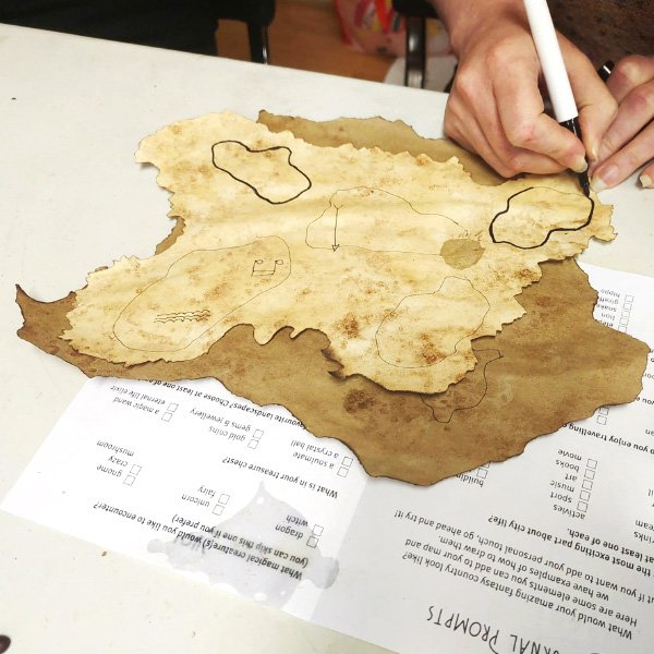 Person drawing landmasses on aged treasure map paper with 'Journal Prompts' worksheet underneath.