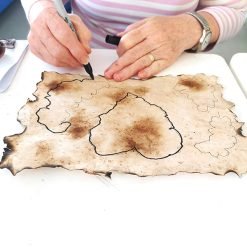 Person tracing shapes on burnt-edged parchment with a black marker, seated at a white table.