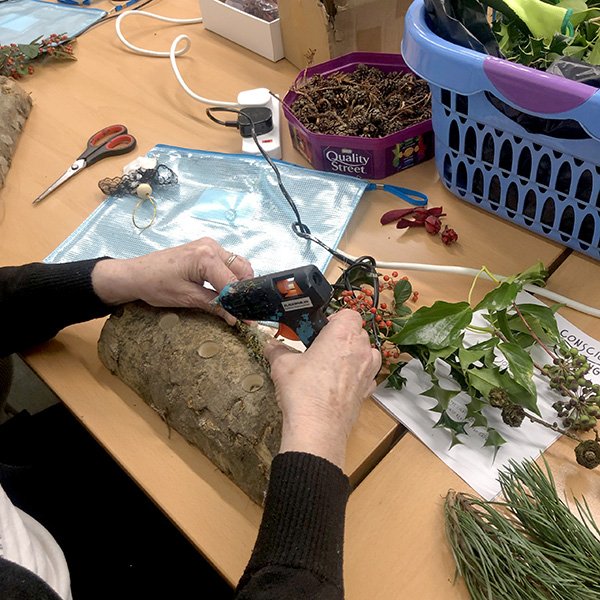 Person gluing red berries and foliage onto tree bark at craft table with pinecones, scissors, and greenery.