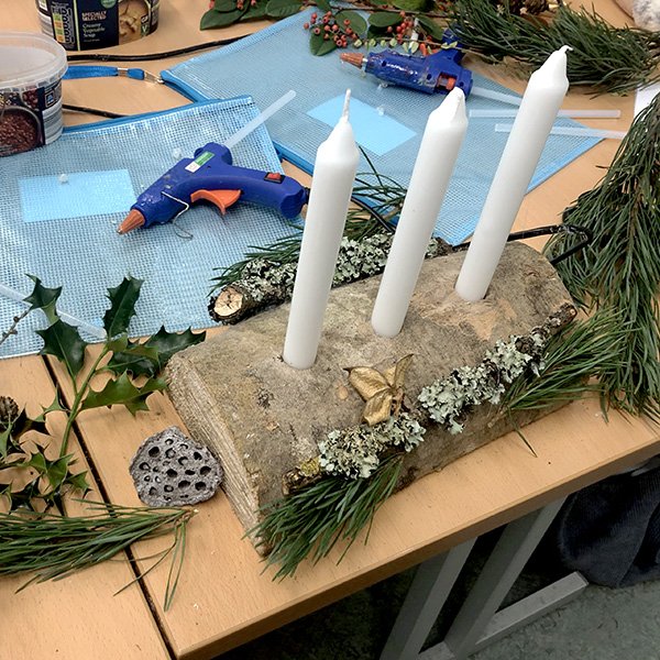 Rustic log candle holder with white candles, pine branches, holly, lotus pod, and golden bow on craft table.