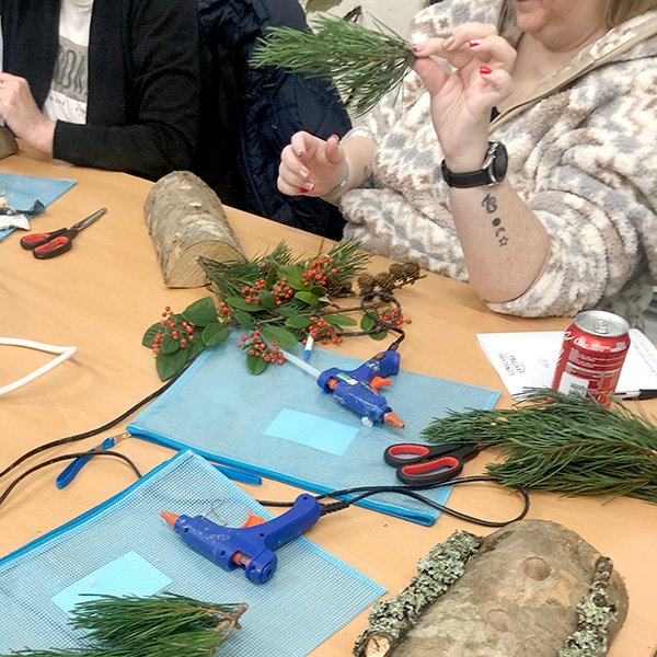 Woman crafting with greenery and pine branches on a table, using hot glue guns; craft supplies, logs, and red berries visible.
