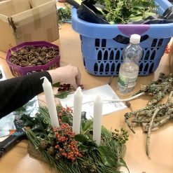 Person at a crafting table arranging a centerpiece with three white candles, evergreen sprigs, and red berries; crafting supplies and a blue laundry basket in the background.