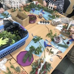 People gathered around a table crafting holiday centerpieces with evergreen branches, pine needles, candles, and red berries; scissors, glue guns, and crafting supplies are scattered across the table.