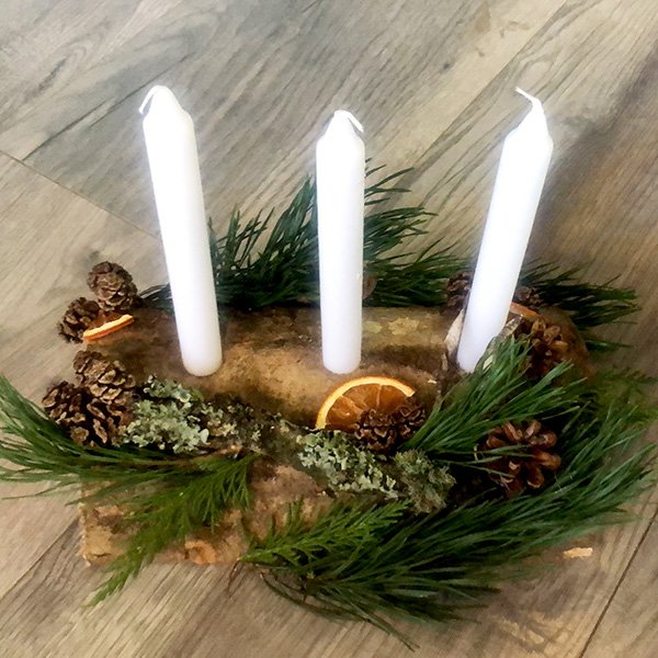 Rustic log centerpiece with three white candles, pinecones, greenery, and dried orange slices on wooden floor.