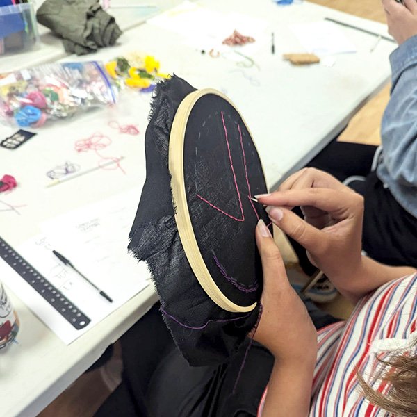 Person stitching pink and purple thread into geometric design on black fabric in embroidery hoop.