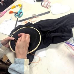 Person stitching orange thread on black fabric in embroidery hoop, with scissors, floss, and notes nearby.