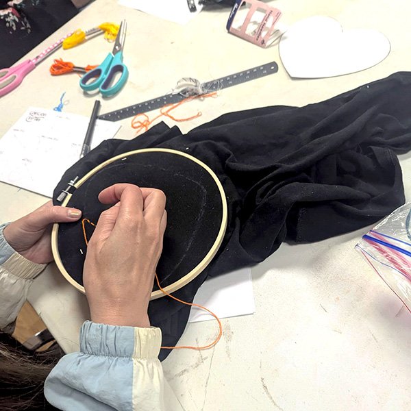 Person stitching orange thread on black fabric in embroidery hoop, with scissors, floss, and notes nearby.