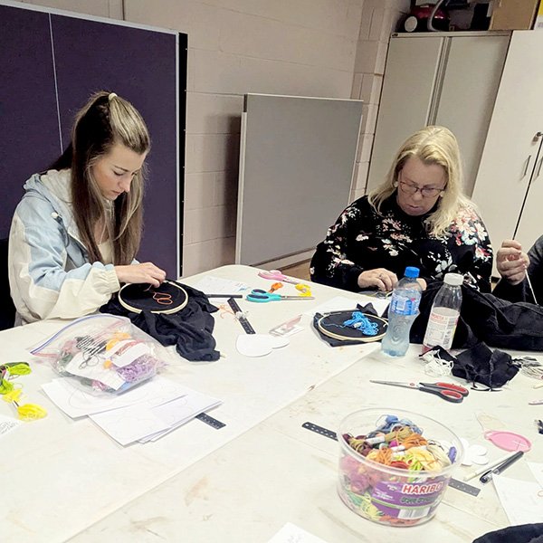 Two people stitching on black fabric in embroidery hoops at a table with threads, tools, and templates.