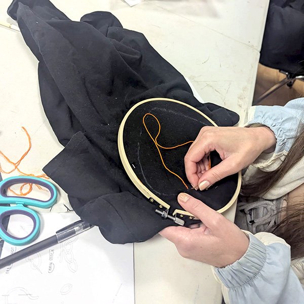Person stitching orange thread on black fabric in embroidery hoop, with scissors and whip stitch guide nearby.