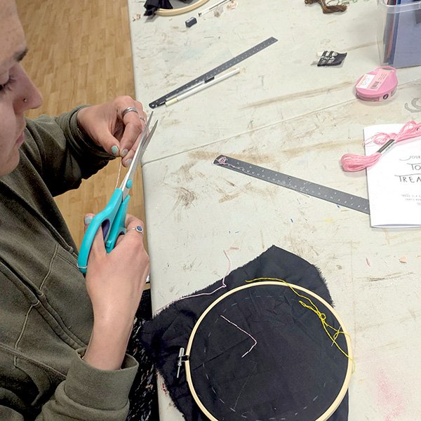 Person cutting thread near embroidery hoop with black fabric, surrounded by floss, tools, and journal booklet.