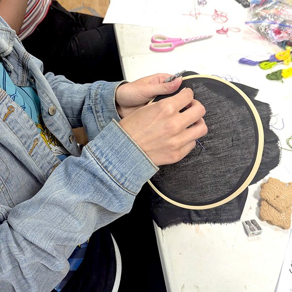 Person stitching purple thread on black fabric in embroidery hoop, with scissors, threads, and craft tools nearby.