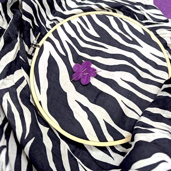 Embroidery hoop holding zebra-print fabric with purple felt flower and two needles inserted.