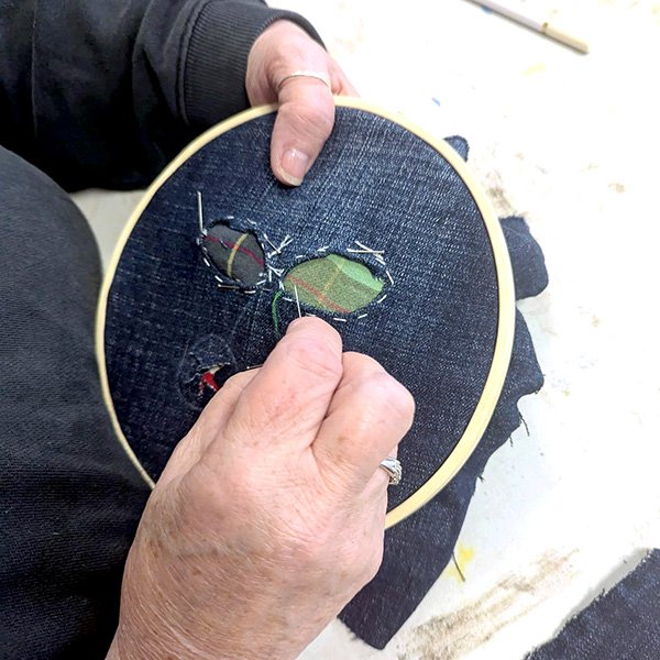 Person stitching around cut-out shapes in denim fabric revealing colorful patterned layer beneath.