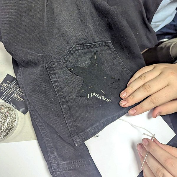 Person sewing black star patch onto black pants pocket with white thread, pins and reference nearby.