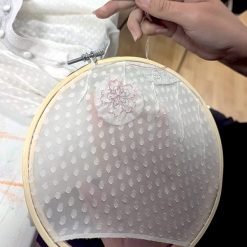 Person stitching pink floral design on sheer polka-dot fabric in embroidery hoop, hand guiding needle.