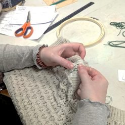 Person stitching green thread onto knitted fabric with scissors, ruler, and embroidery tools nearby.