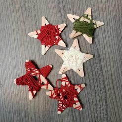Five star-shaped ornaments made from wooden craft sticks wrapped with yarn in red, white, and green; each star features crisscrossed yarn patterns and is arranged on a wooden surface, showcasing a festive DIY holiday decoration project