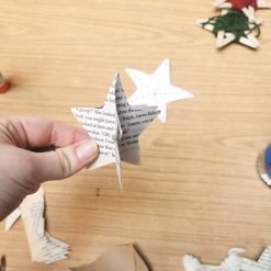 Hand holding a three-dimensional star ornament made from interlocked paper cutouts with printed text; background shows additional star-shaped paper pieces, including one decorated with green yarn and a red center, all arranged on a wooden table