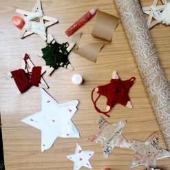 Holiday-themed craft project on a wooden table featuring star-shaped ornaments made from popsicle sticks wrapped in red, green, and white yarn; includes paper stars with printed holiday text and red dots, a glue stick, red glitter glue, two cardboard rolls, and a roll of festive wrapping paper