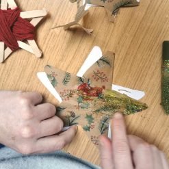 Person decorating a star-shaped cutout made from patterned paper with holly leaves, berries, and pine branches; applying red and yellow glitter using a green glitter glue tube; nearby are additional paper stars and a popsicle stick ornament wrapped in red yarn, all arranged on a wooden surface