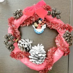 Festive Christmas wreath hanging on a dark wooden surface, wrapped in red fabric and decorated with red tinsel garland, pinecones (some painted white), a red bow at the top, and a central ornament featuring a figure riding a blue and white polka-dotted rocking horse