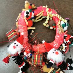 Handmade Christmas wreath placed on a dark wooden surface, wrapped in red material and decorated with plush Santa Claus faces, miniature plaid gift boxes, colorful pom-poms, felt ornaments, small clothespins, and curled yellow ribbon