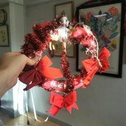 Hand holding a handmade Christmas wreath decorated with red tinsel garland, four evenly spaced red bows, and a small figure of a reindeer pulling a sleigh at the top center; visible tape and string secure the decorations; background includes framed artwork and sunlight streaming into the room