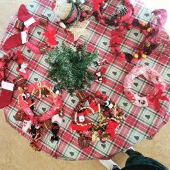 Round table with red and white plaid tablecloth featuring green Christmas tree patterns, displaying handmade Christmas decorations including festive wreaths with red ribbons, pinecones, miniature gift boxes, snowmen, penguins, small stockings, and a central artificial green Christmas tree arrangement