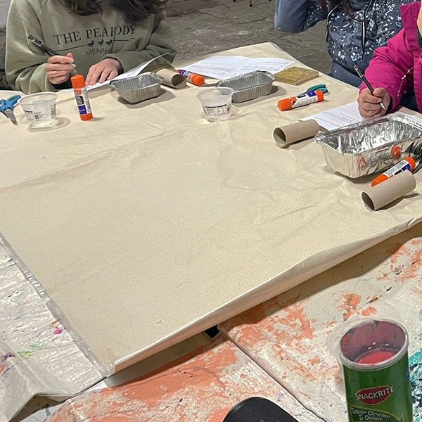 Group of individuals seated around a beige-covered table engaged in a craft or educational activity; supplies include glue sticks, paper towel rolls, aluminum trays, plastic cups with water, and sheets of paper; one person wears a sweatshirt reading 'THE PEABODY MEMPHIS' as participants write or draw on paper in a classroom or community center setting