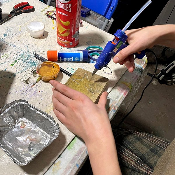Person using a blue hot glue gun to apply glue onto a small wooden rectangle with a star outline; tabletop is cluttered with crafting supplies including scissors, a glue stick, black marker, yellow paint or adhesive, a Pringles can, and a paint-stained aluminum tray