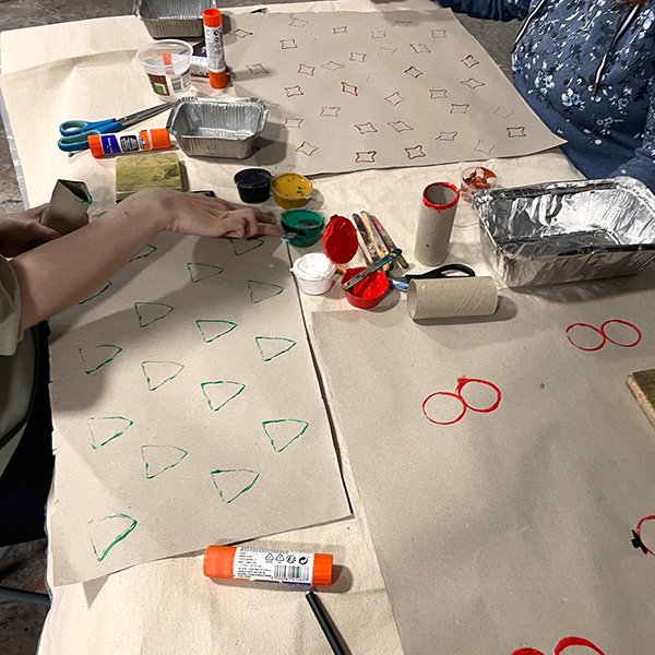 Two people stamping shapes onto large sheets of paper using cardboard tubes and paint; stamped shapes include green triangles, red circles, and red star-like forms arranged in a grid pattern; tabletop contains paint containers in black, yellow, green, red, and white, glue sticks, scissors, paintbrushes, and aluminum trays