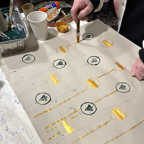 Person painting golden stripes onto circular ornaments printed on a large sheet of paper; each ornament features a green Christmas tree inside a black circle, arranged in a grid with vertical lines resembling hanging decorations; tabletop includes paint containers, brushes, glue sticks, and cups