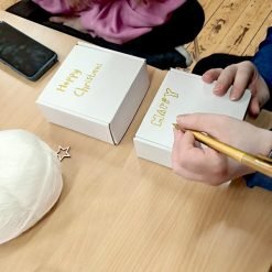 Person using a gold pen to write 'HAPPY' on a white cardboard box, with another box labeled 'Happy Christmas' nearby; tabletop includes white yarn, a wooden star ornament, a smartphone, and people seated around the table, suggesting a festive craft or gift-wrapping activity