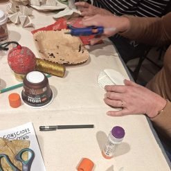 Two people engaged in a holiday-themed craft activity at a table, one using a hot glue gun to assemble folded paper decorations; tabletop includes a red glittered ornament, gold glitter tube, glue stick, black pen, scissors, a container labeled 'HIGH PROTEIN', a paint-stained paper plate, and red patterned paper; a handwritten note reading 'CONSCIOUS CRAFTING' is visible