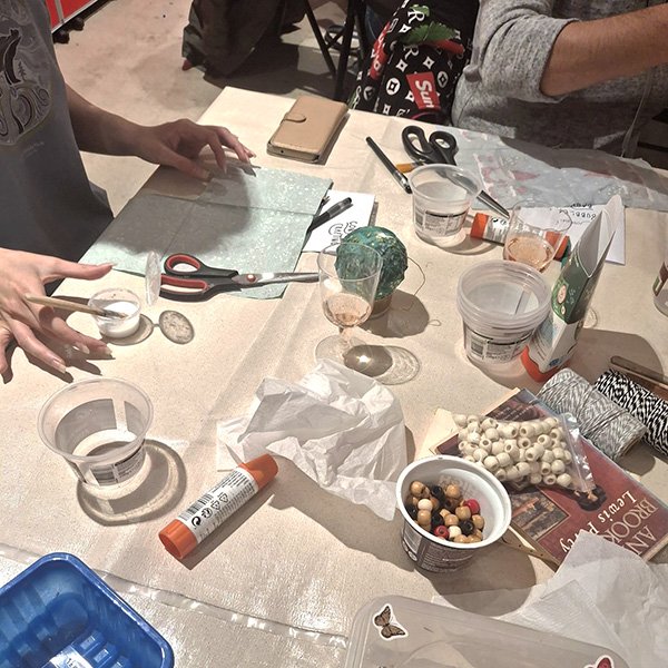 Crafting workspace with multiple people engaged in a DIY project; table covered with supplies including glue sticks, scissors, plastic containers, string, wooden and plastic beads, tissue paper, a green decorative ball, patterned paper, books, and a smartphone; one person using a spoon and small container, possibly mixing or applying a substance