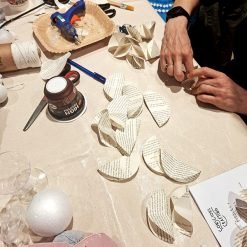 Person engaged in a paper craft activity using semicircular book pages; tabletop includes a hot glue gun, scissors, pen, spool of string, foam ball, and a container labeled 'HIGH PROTEIN'; visible text on paper reads 'CHAPTER' and 'COLD COUSINS', suggesting creative reuse of printed materials